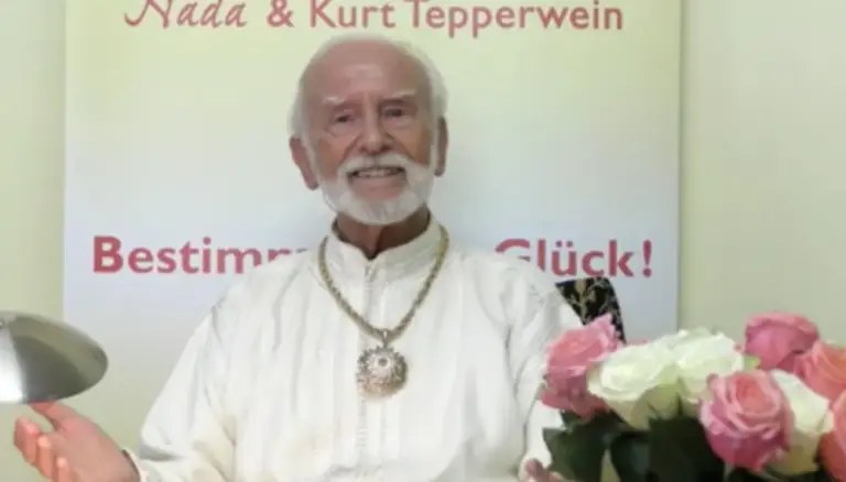 Kurt Tepperwein in weißem Gewand vor einem Banner, rechts ein Rosenstrauß