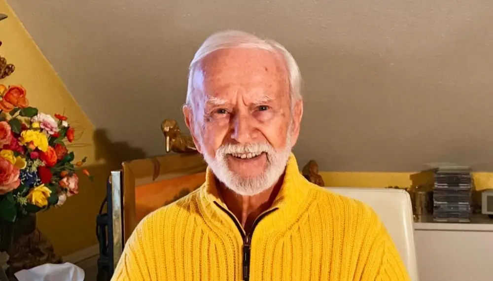 Kurt Tepperwein in gelbem Pullover lächelt, im Hintergrund Blumen