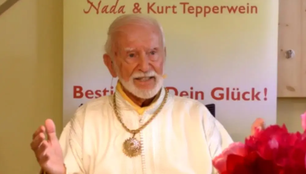 Kurt Tepperwein in weißem Gewand gestikuliert vor einem Banner mit der Aufschrift „Nada & Kurt Tepperwein“