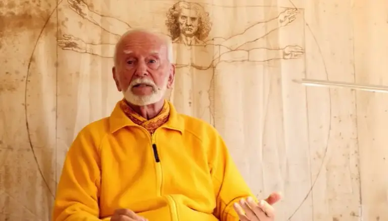 Kurt Tepperwein in gelber Jacke spricht vor einer Holzwand mit Zeichnung