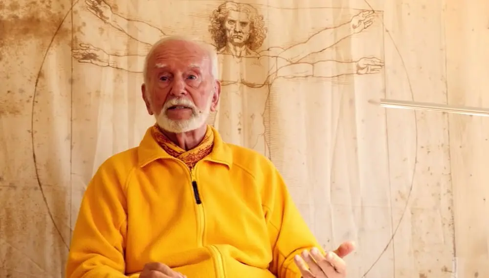 Kurt Tepperwein in gelber Jacke spricht vor einer Holzwand mit Zeichnung