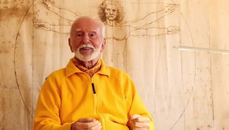 Kurt Tepperwein in gelber Jacke spricht vor einer Holzwand mit Zeichnung