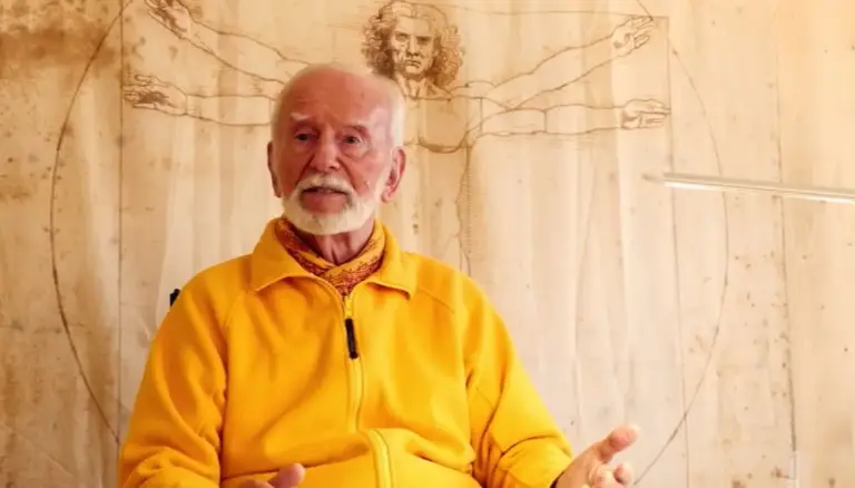 Kurt Tepperwein in gelber Jacke spricht vor einer Holzwand mit Zeichnung