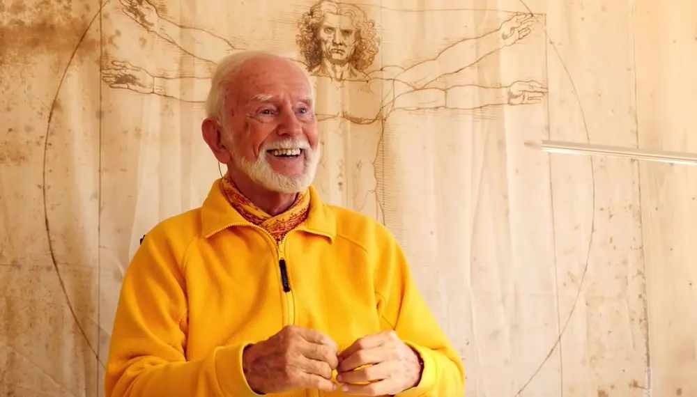 Kurt Tepperwein in gelber Jacke lächelt vor einer Holzwand mit Zeichnung