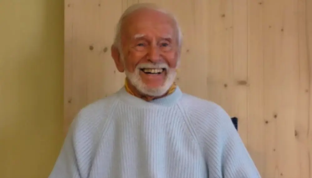 Kurt Tepperwein in hellem Strickpullover lacht vor einer Holzwand