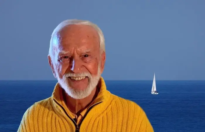 Kurt Tepperwein in gelbem Pullover lächelt vor blauem Meer mit Segelboot
