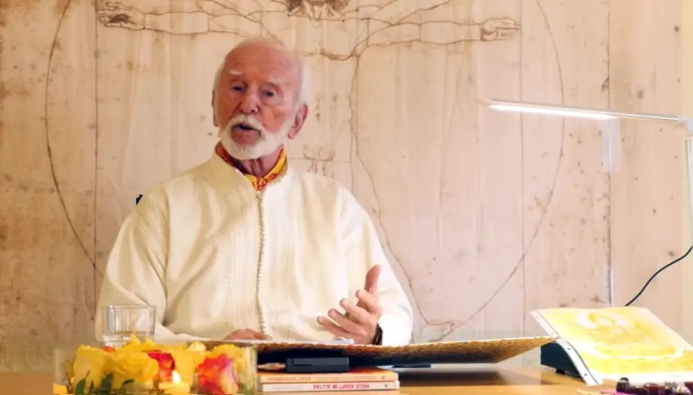 Kurt Tepperwein in weißem Gewand spricht am Tisch vor einer Holzwand mit Zeichnung