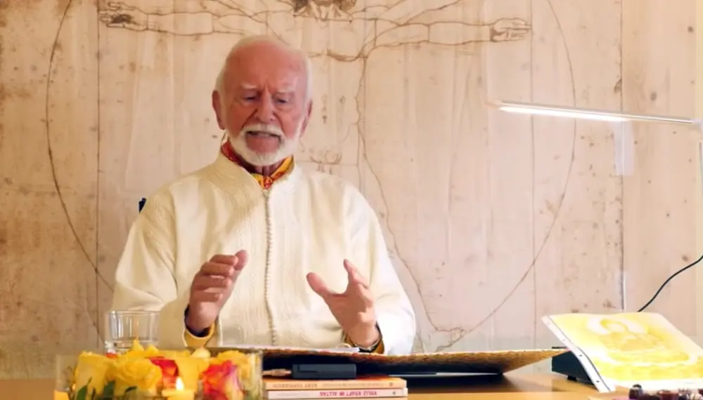 Kurt Tepperwein in weißem Gewand spricht am Tisch vor einer Holzwand mit Zeichnung