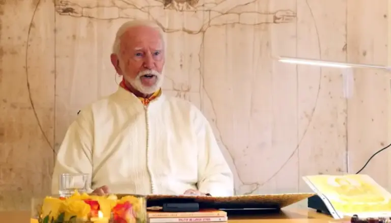 Kurt Tepperwein in weißem Gewand sitzt lächelnd am Tisch vor einer Holzwand