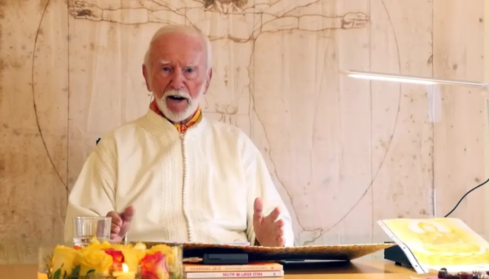 Kurt Tepperwein in weißem Gewand spricht am Tisch vor einer Holzwand