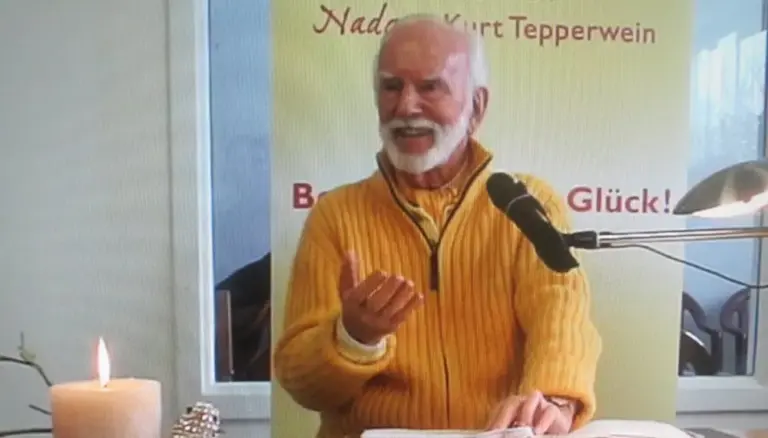 Kurt Tepperwein in gelbem Pullover spricht am Mikrofon vor einem Banner, vorne eine brennende Kerze
