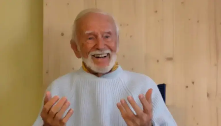 Kurt Tepperwein in hellem Strickpullover gestikuliert lächelnd vor einer Holzwand