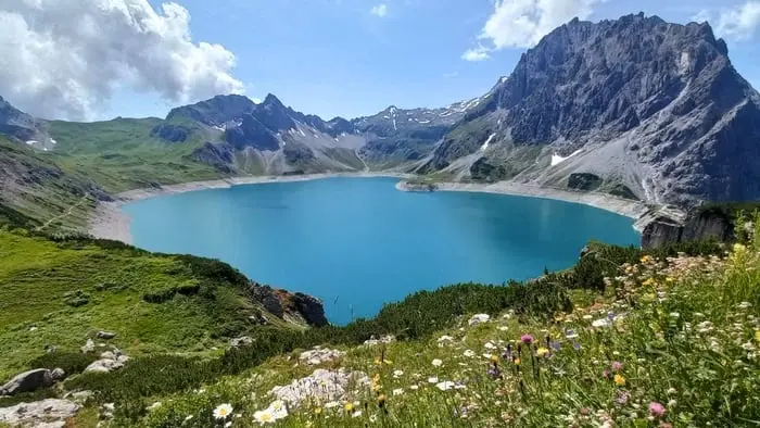 Blick von oben auf den türkisblauen Lünersee mit Bergpanorama und Blumenwiese